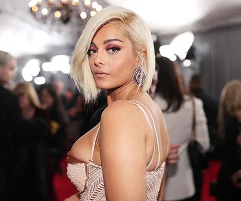 A woman with blonde hair and pink dress posing on a red carpet, with chandelier and crowd in the background.