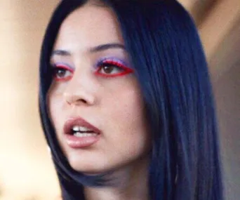 A woman with sleek black hair wears bold makeup with red eyeliner and purple eyeshadow, mouth slightly open.