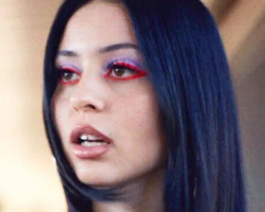 A woman with sleek black hair wears bold makeup with red eyeliner and purple eyeshadow, mouth slightly open.