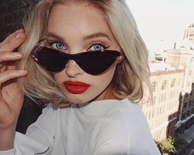 Woman with blonde hair, red lipstick, and cat-eye sunglasses, posing outdoors with buildings in the background.