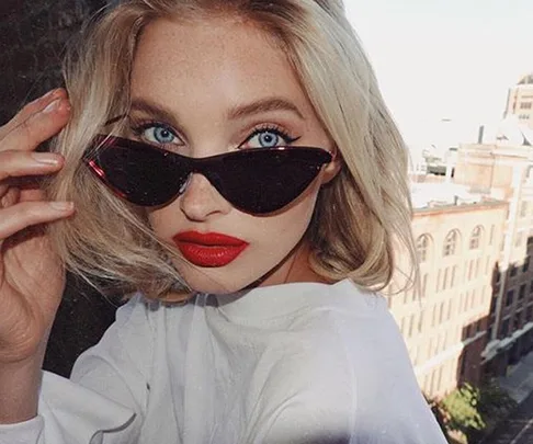 Woman with blonde hair, red lipstick, and cat-eye sunglasses, posing outdoors with buildings in the background.