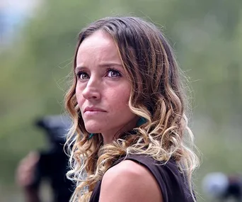 A woman with long curly hair looks off-camera, appearing concerned, with blurred greenery in the background.