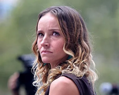 A woman with long curly hair looks off-camera, appearing concerned, with blurred greenery in the background.