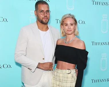 A couple poses at a Tiffany & Co. event; the man wears glasses and a beige suit, the woman wears a black top and plaid skirt.