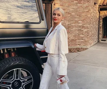 Woman in a white suit standing by a luxury SUV, holding a clear handbag, with a brick building in the background.