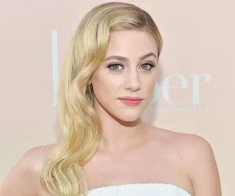 A woman with long, wavy blonde hair and a white strapless top poses against a soft-colored background.