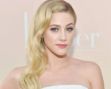 A woman with long, wavy blonde hair and a white strapless top poses against a soft-colored background.