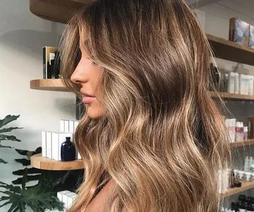 Woman with long, wavy, caramel-highlighted hair in a salon setting.