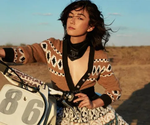 Woman in patterned cardigan posed by motorcycle in desert landscape.