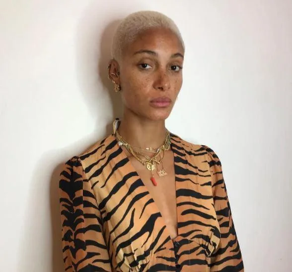 A person with short blonde hair, wearing a tiger print top and layered necklaces, stands against a plain background.