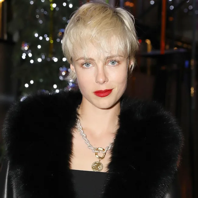 Young woman with short blonde hair, red lipstick, wearing a fur collar and necklace with a medallion, in festive setting.