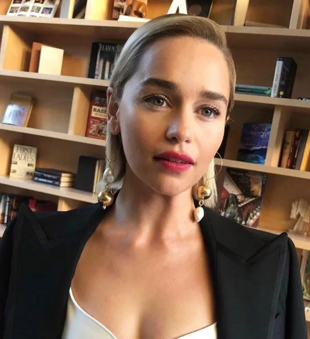 A woman with earrings and a black outfit stands in front of a bookshelf.