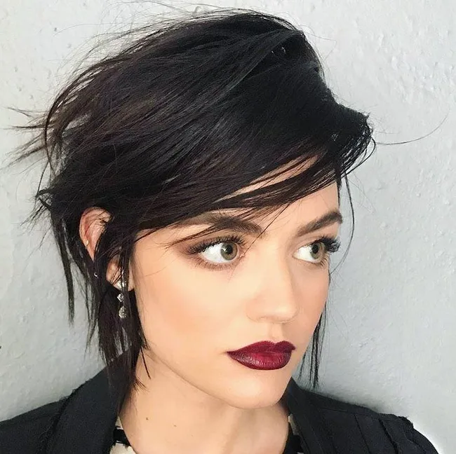 Short-haired person with dark, tousled hair, dramatic makeup, burgundy lipstick, and earrings, looking to the side.