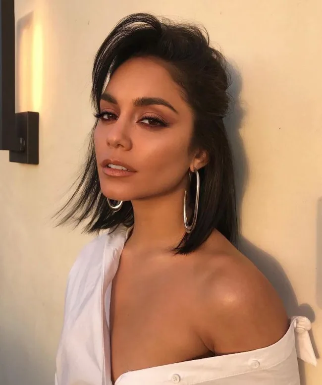 A woman with shoulder-length hair, wearing hoop earrings and an off-shoulder white top, stands against a beige wall.