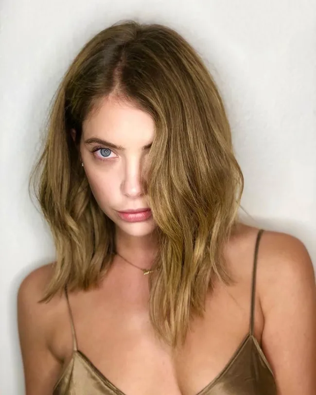 Woman with wavy shoulder-length hair, wearing a gold camisole, looking directly at the camera against a neutral background.