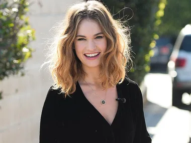 A person with wavy hair wearing a black top, smiling outdoors.