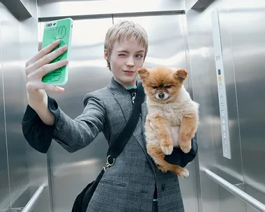 Person winking, taking a selfie in an elevator with a fluffy dog in their arm.