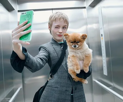 Person winking, taking a selfie in an elevator with a fluffy dog in their arm.