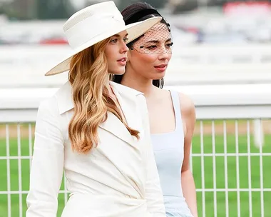 Two women dressed in elegant race day outfits, one in a white suit with a hat, the other in a powder blue dress with a fascinator.