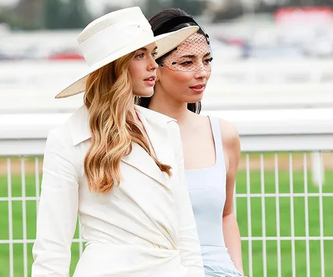 Two women dressed in elegant race day outfits, one in a white suit with a hat, the other in a powder blue dress with a fascinator.