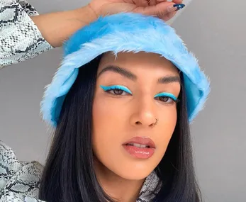 Person with blue eyeliner and hat poses confidently against a neutral background.