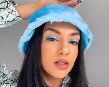 Person with blue eyeliner and hat poses confidently against a neutral background.