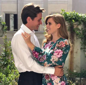 A couple embracing, wearing a floral dress and a white shirt, in a garden setting. Both are smiling at each other.