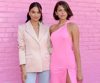 Two women posing in front of a pink brick wall; one in a beige blazer and jeans, the other in a pink one-shoulder dress.