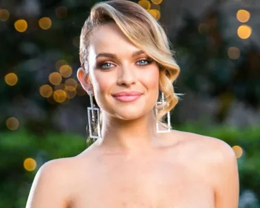 A woman with short blonde hair and statement earrings smiling in an outdoor setting with blurred lights in the background.