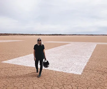 Person walking on a dry lakebed in the desert, holding a helmet, with large white markings on the ground.