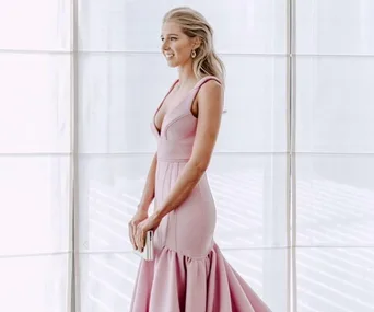 Woman in a light pink gown with a deep neckline, holding a clutch, standing near a window with sheer curtains.