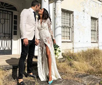 Couple kissing in front of an old building, bride in lace dress with bouquet, groom in light blazer.