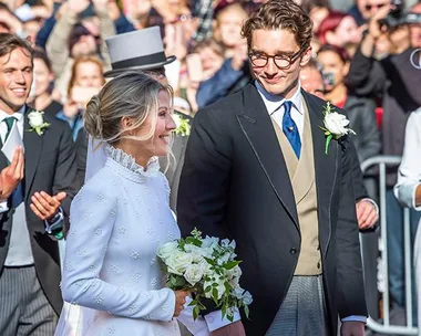 Bride in a white dress holding a bouquet, smiling at groom in a black suit, surrounded by guests.