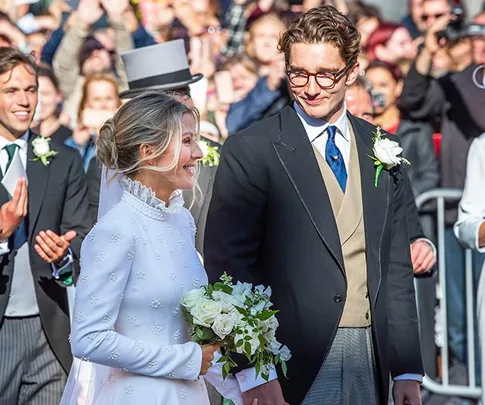 Bride in a white dress holding a bouquet, smiling at groom in a black suit, surrounded by guests.
