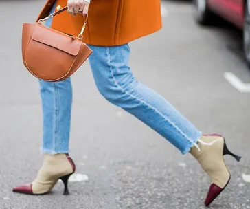 Woman in orange coat with brown handbag, blue jeans, and two-tone heels walking on street.