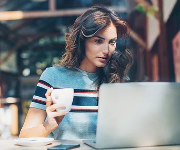 Woman with long hair holding a coffee cup, focused on a laptop screen in a bright, modern setting.