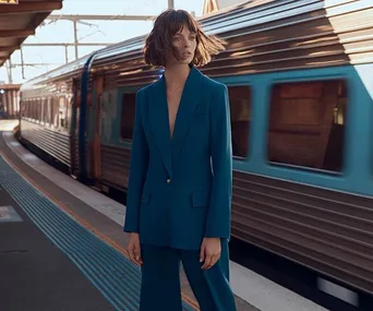 Person in teal suit standing on train platform, hair blowing, with train passing in the background.