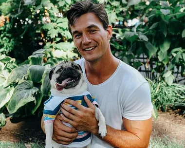 Man in a white shirt smiling, holding a pug in a colorful outfit, with greenery in the background.