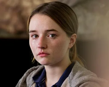 A young woman with a solemn expression, wearing a grey hoodie, looks at the camera with teary eyes.