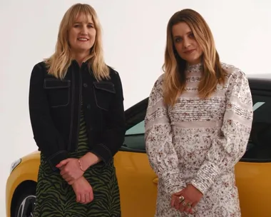 Two women stand in front of a yellow car, one wearing a black jacket and the other in a floral dress.