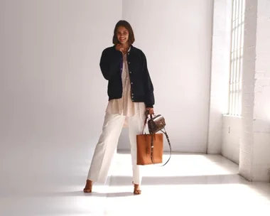 Woman in a navy jacket and white pants, holding two bags, stands smiling in a brightly lit room.