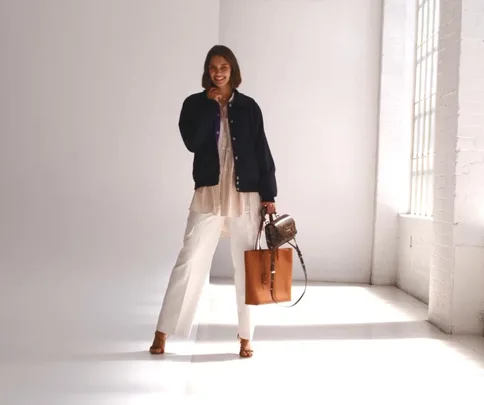 Woman in a navy jacket and white pants, holding two bags, stands smiling in a brightly lit room.