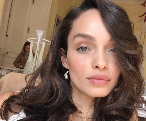 A woman with long, wavy brown hair takes a selfie in a room, another person in the background.