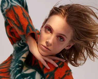 Model poses in a vibrant orange and green sweater, with dynamic hair and bold eye makeup, creating an artistic look.