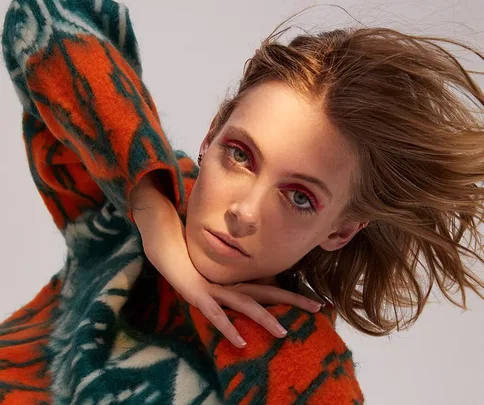 Model poses in a vibrant orange and green sweater, with dynamic hair and bold eye makeup, creating an artistic look.