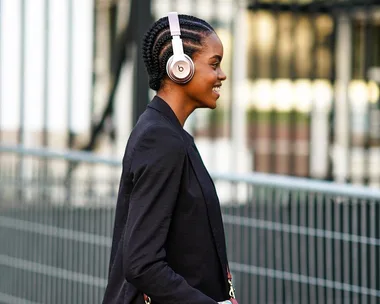 Person walking outdoors wearing headphones, smiling, with braided hair and a black blazer.