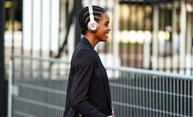 Person walking outdoors wearing headphones, smiling, with braided hair and a black blazer.