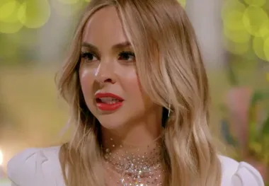 Woman with long blonde hair and red lips, wearing a sparkly necklace and white outfit, with a blurred green background.