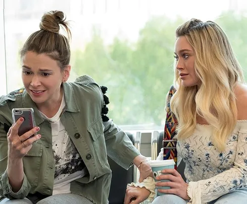 Two women sit together; one looks concerned at her phone, while the other listens, holding a cup.