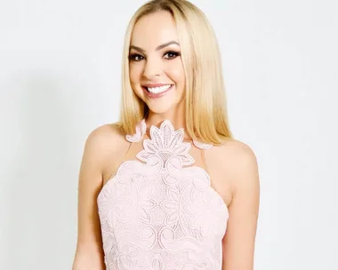 A woman in a white lace dress, smiling with long blonde hair against a plain background.